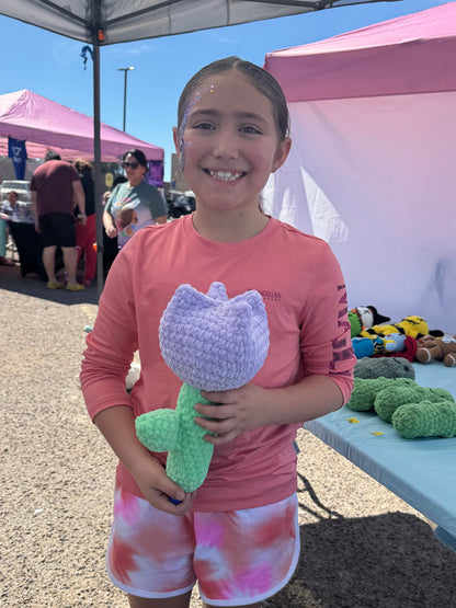 Child holding a crochet lavender tulip in front of a pink tent with various items on display.